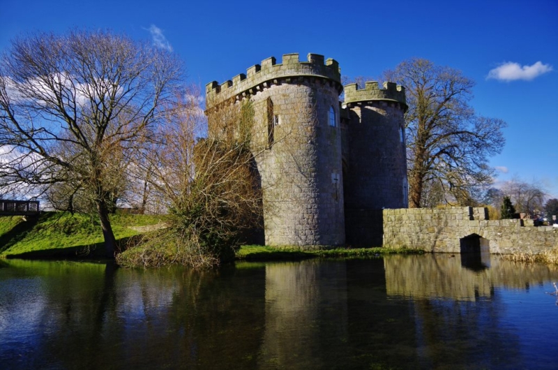 Whittington Castle on a sunny day in front of a lake. You can see a reflection of the old castle in the lake.